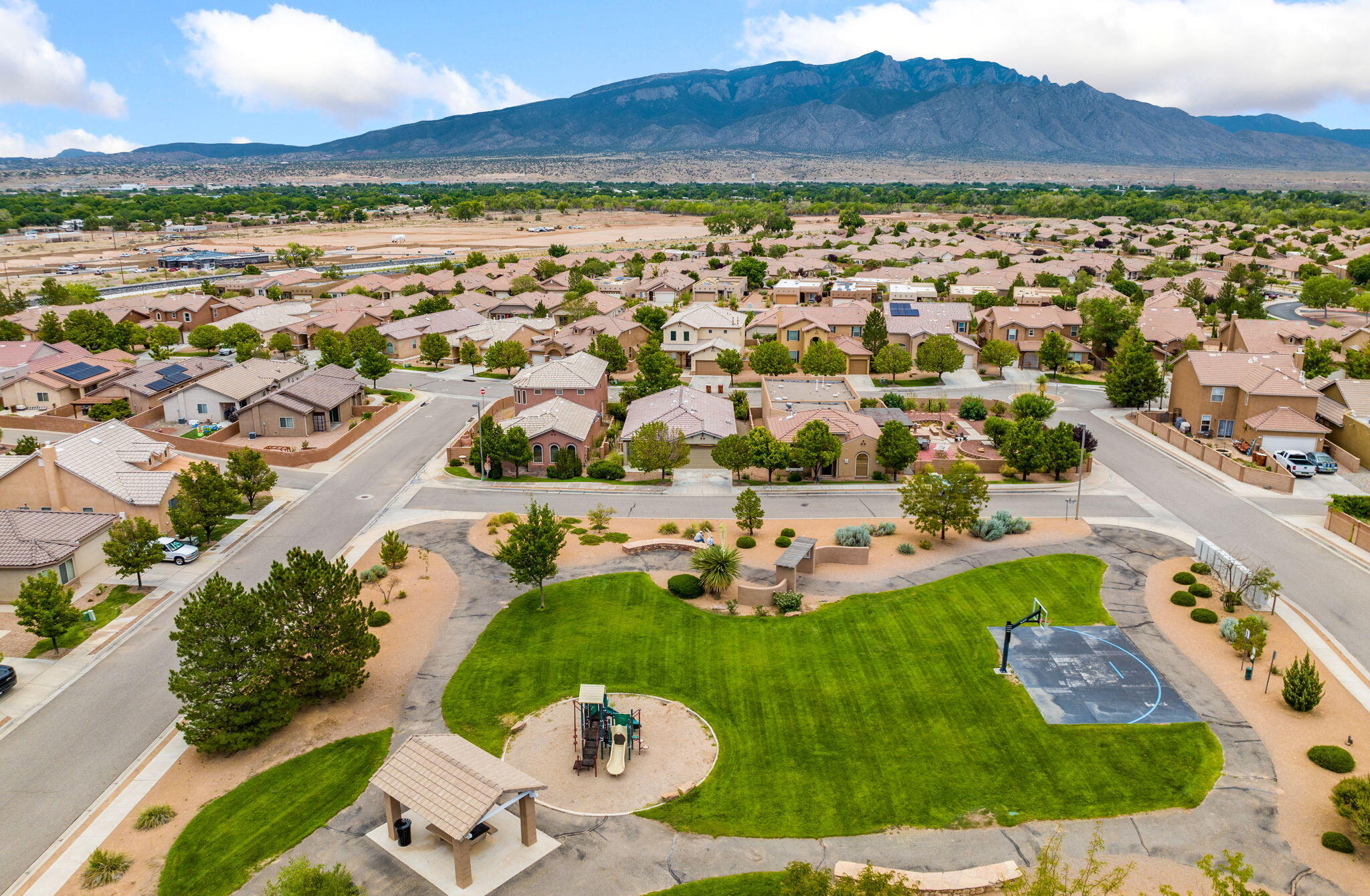 Aerial view from 806 Vargas Drive toward the grassy neighborhood park, walking paths, play area, basketball court, and Sandia Mountains beyond.