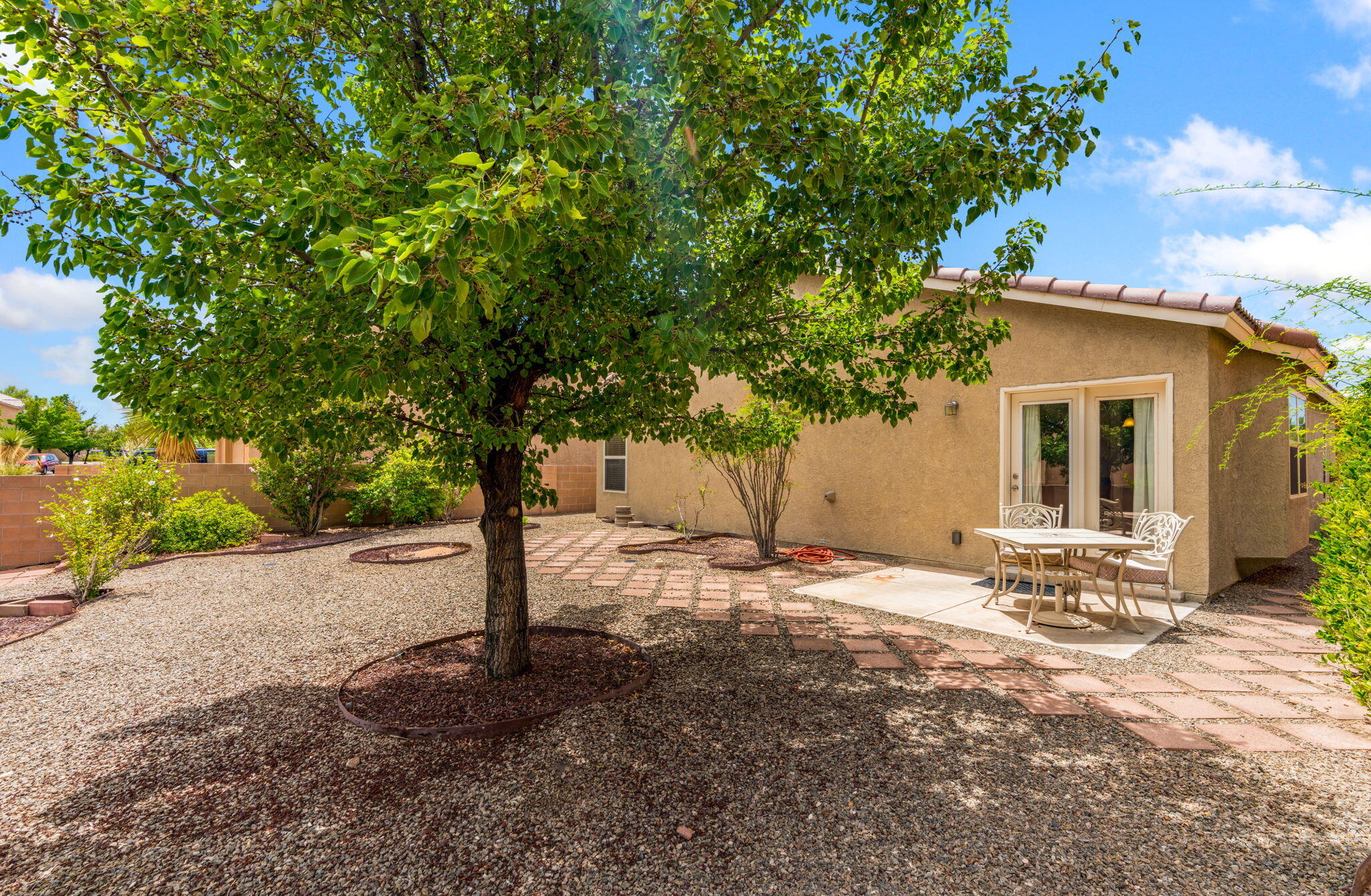 Landscaped backyard at 806 Vargas Drive with a mature shade tree, gravel, planting beds, patterned patio areas, and an outdoor table beside the home.