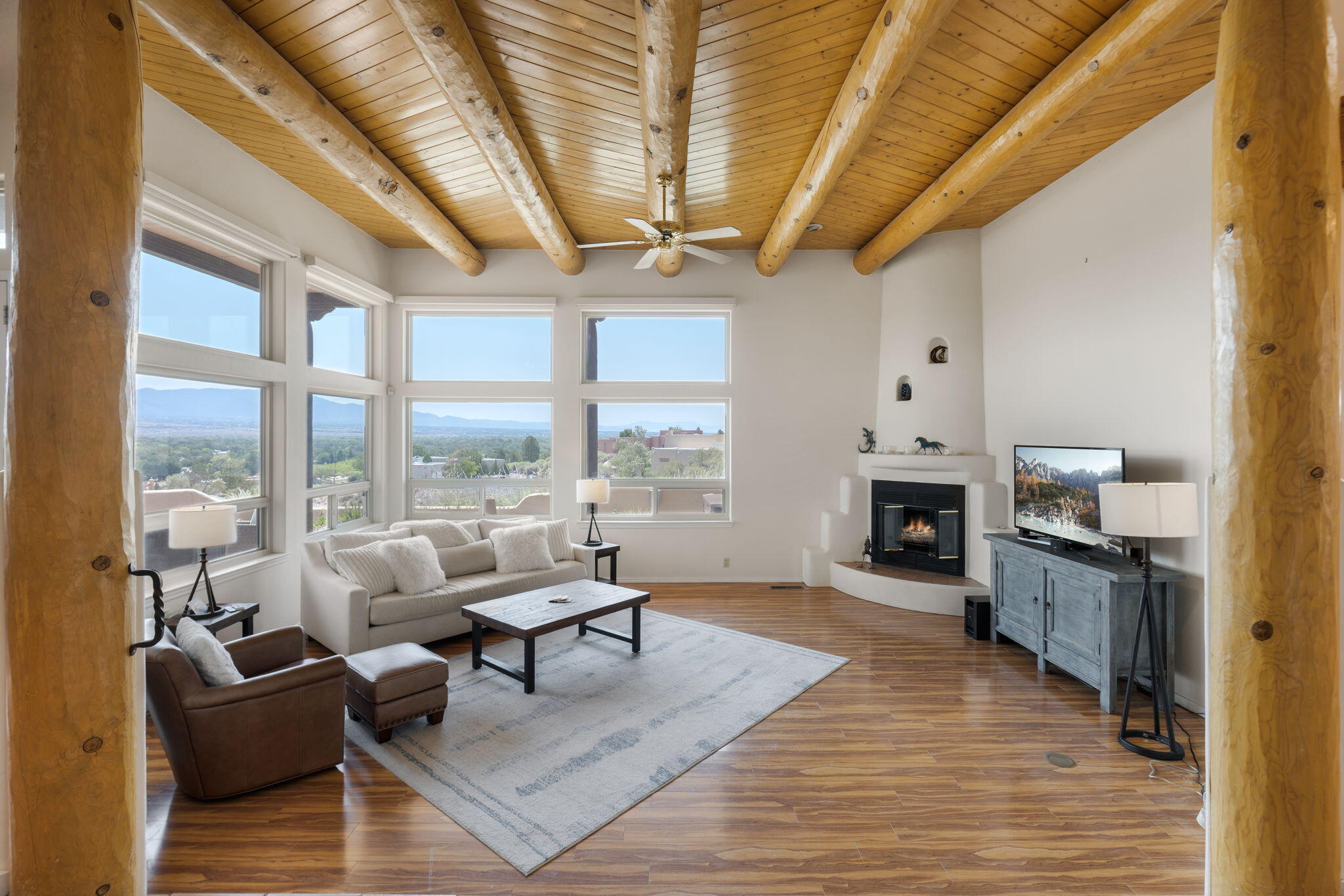 Great room at 40 Sereno Road in Corrales with large vigas, a Kiva fireplace, and broad windows framing mountain and valley views