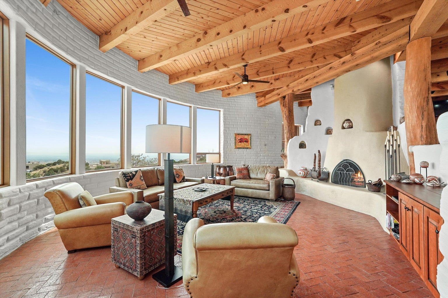 Living room with a wood-burning kiva fireplace, brick floors, wood beams, and city-facing windows